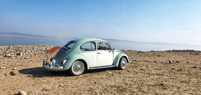 Pale Volkswagen Beetle with a raised red-slatted deck-lid rack beside a rocky shoreline