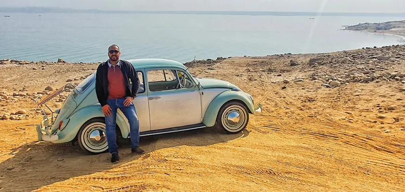 Person standing beside a pale Volkswagen Beetle with a raised deck-lid rack near the water